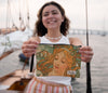 A person holding a wristlet bag featuring a printed artwork of a woman's portrait in warm tones with flowing lines.